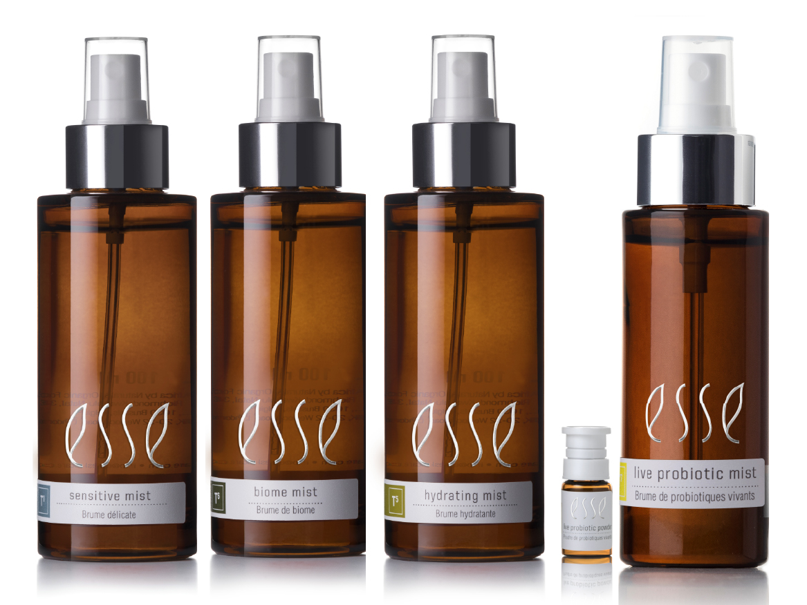 Four brown spray bottles labeled "Esse" (sensitive mist, biome mist, hydrating mist, and live probiotic mist) are lined up side by side, with a small clear vial in front of the third bottle, against a white background.