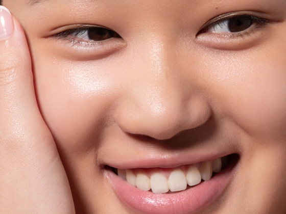 A close-up of a smiling person with clear, smooth skin, showing their teeth and part of their hand gently touching their face.